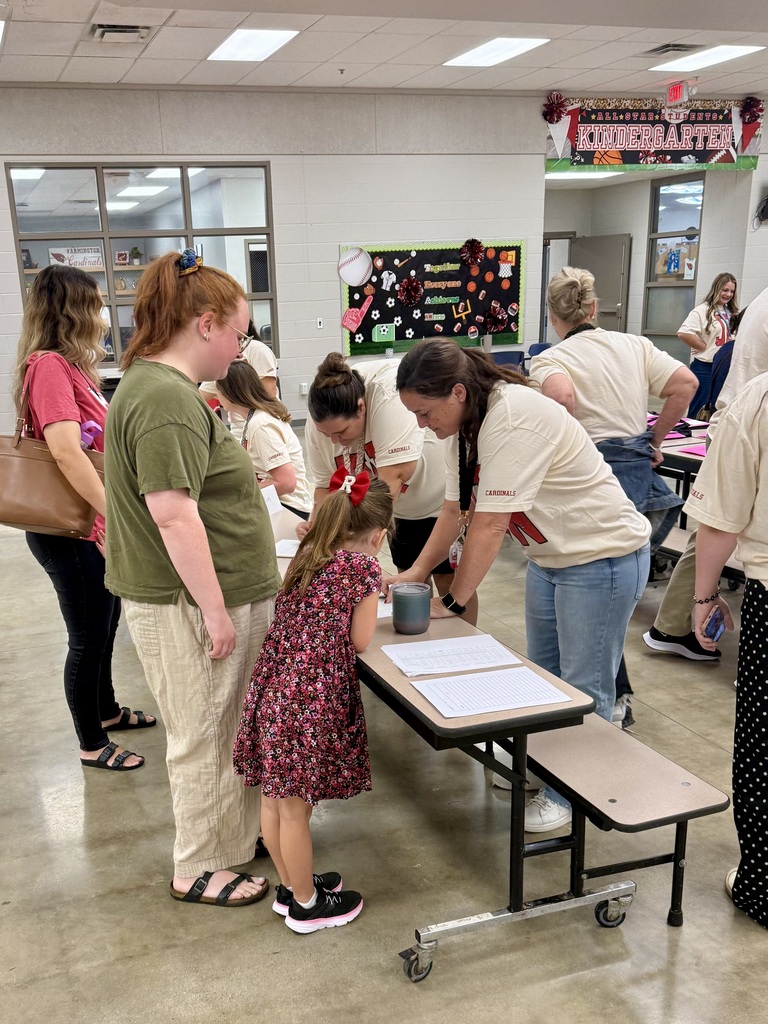 Adults and children are standing around tables, some bending over to look at papers while others talk.