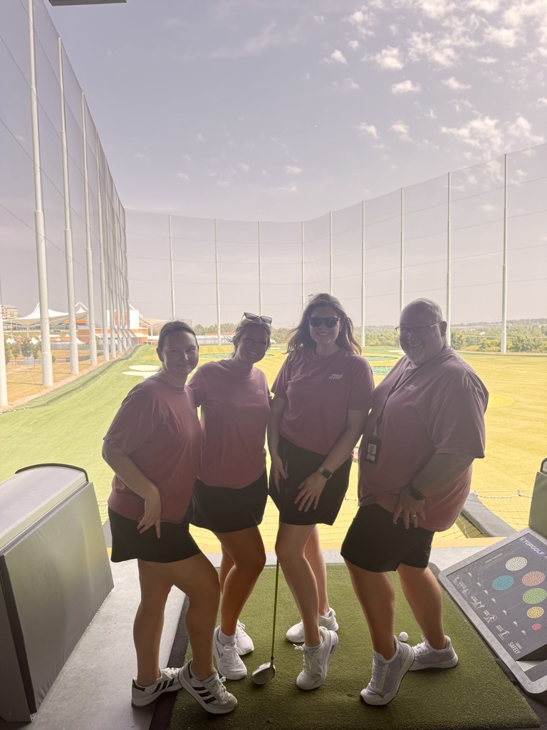 Four people wear matching shirts and shorts. They stand on a green mat inside a golf practice facility.