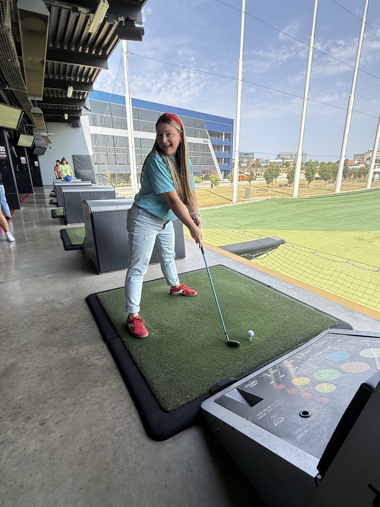 A young person stands on a green mat and swings a golf club at an indoor driving range.