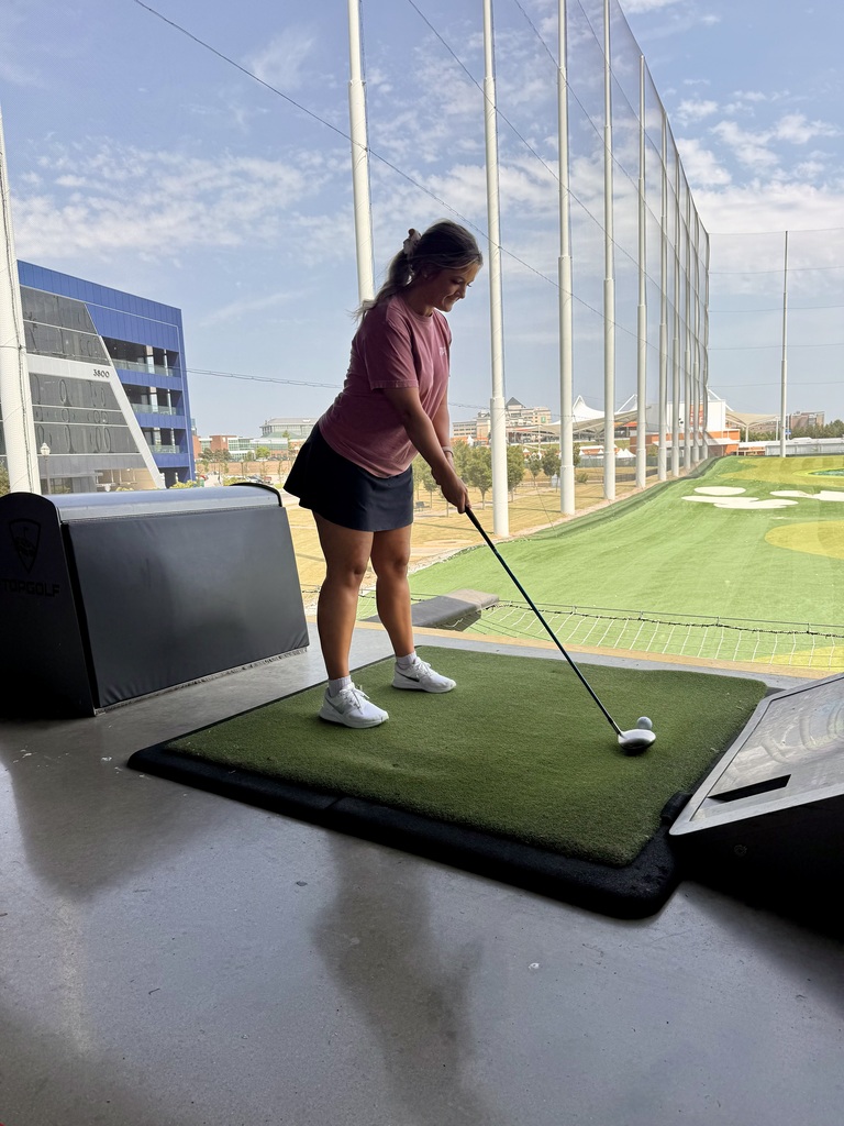 A woman practices golf indoors. She stands on a green mat, holding a club. The area is enclosed by glass walls.