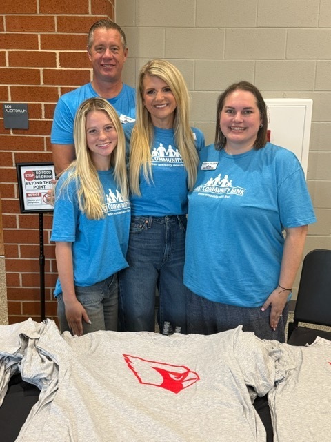 Four adults in blue t-shirts stand behind a table with white shirts displayed. One adult stands on the left, two in the middle, and one on the right. A brick wall is in the background.