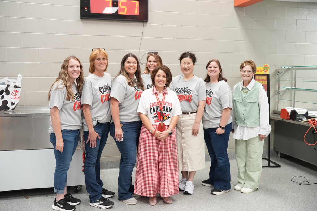 Nine women in matching shirts stand in a room with a clock on the wall and a table with a cow cutout.