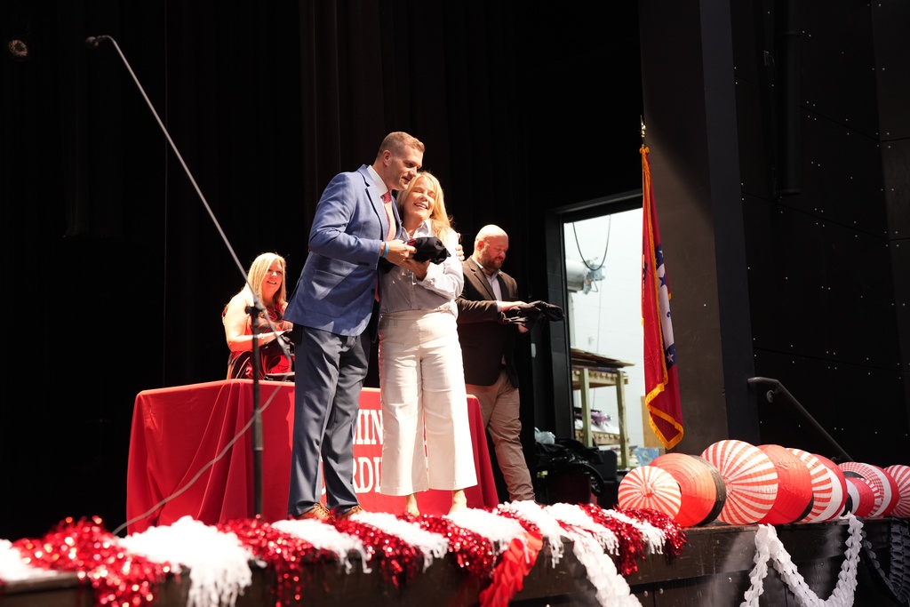 A stage with a red table in the background. A man and a woman are on stage holding a camera.