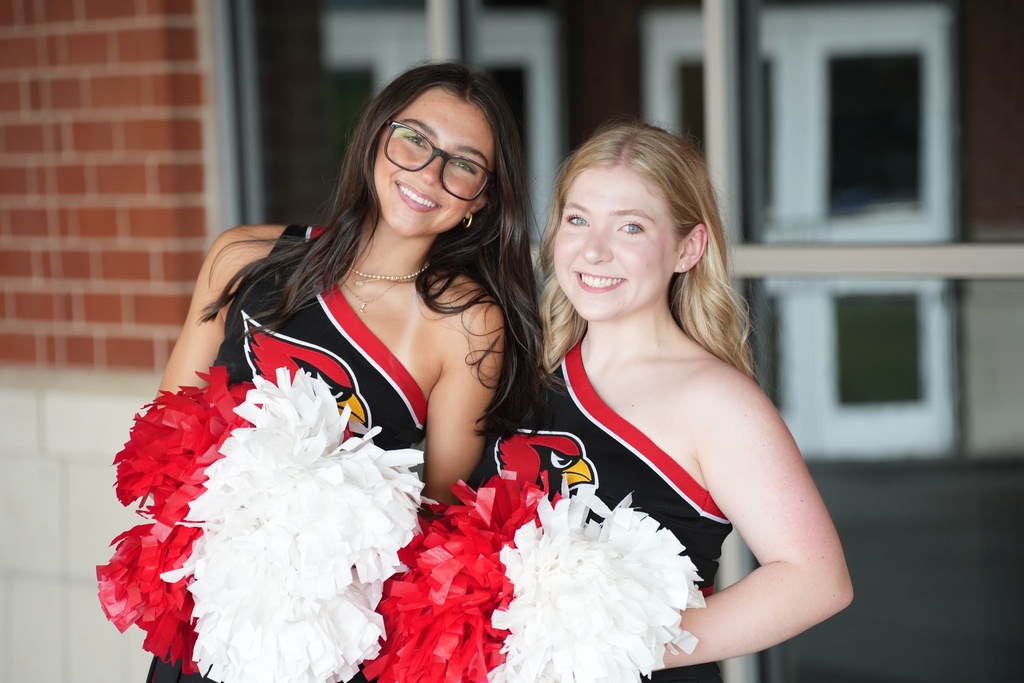 Two women in cheerleading uniforms holding red and white pom-poms stand in front of a brick wall.