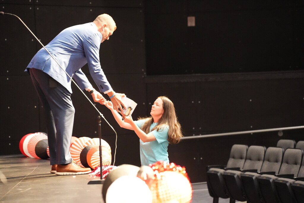 A man in a blue suit hands an item to a girl in a blue shirt on stage.
