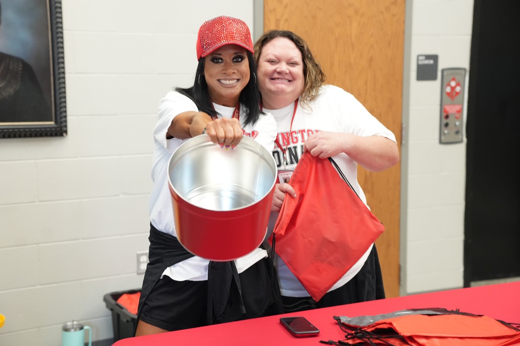 Two women in white t-shirts and black shorts, one holding a red bucket, smile for a photo in a room with a red table and framed picture.