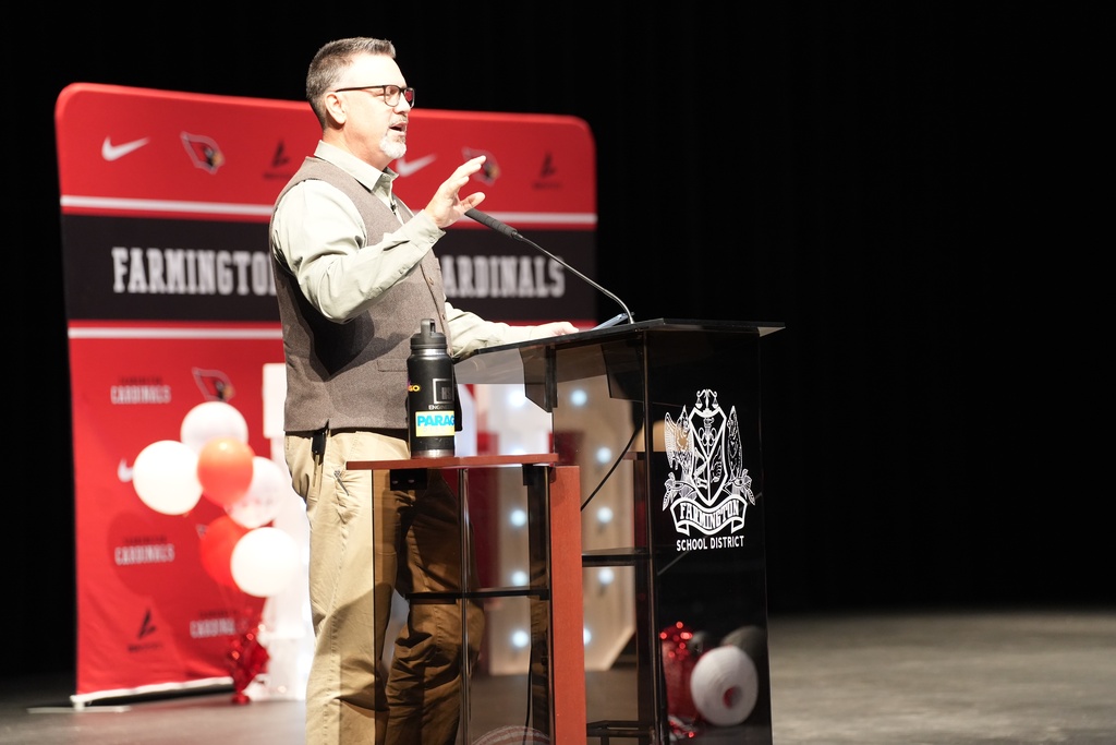 A man speaking into a microphone at a podium, with a red banner behind him featuring text and logos.