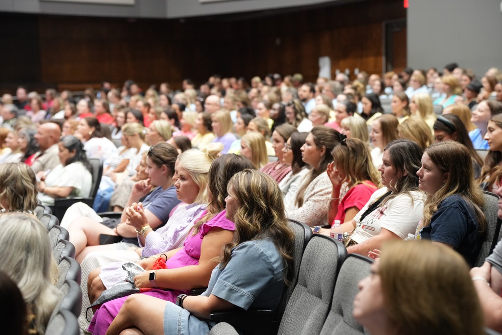 Large group of people seated in chairs inside a large room. Some are looking forward, others are engaged in conversation.