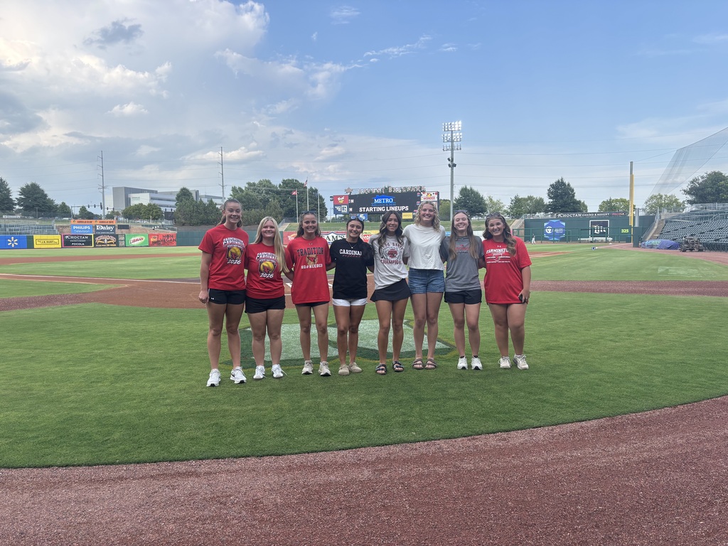 A group of people, all wearing red shirts, stand on a baseball field with a blue sky and clouds in the background.