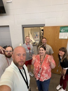 Five adults stand in a room, smiling and making hand gestures. One man holds a camera. Behind them, there are doors and a wall with a bulletin board.