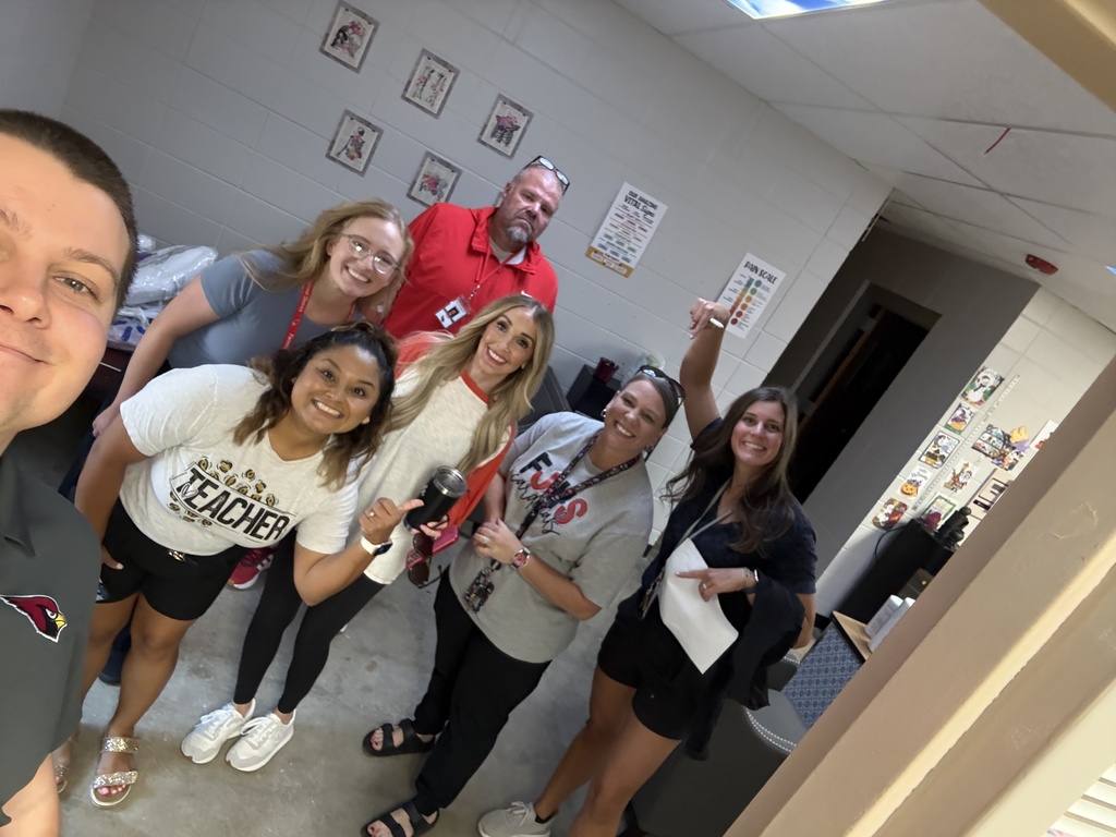 A group of adults in matching white t-shirts stands in a hallway. Two men and five women smile for the camera.