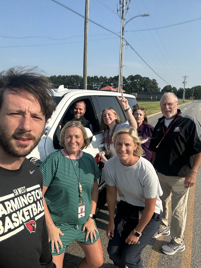 Six people stand in front of a white SUV. The man on the left wears a black t-shirt. The two women in the middle wear green and white clothes. The man on the right wears a black jacket.