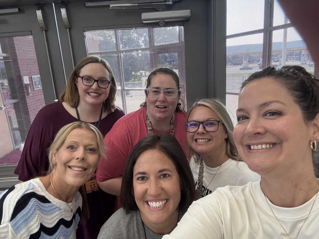 Six women smile for a photo near a door with large windows in the background.