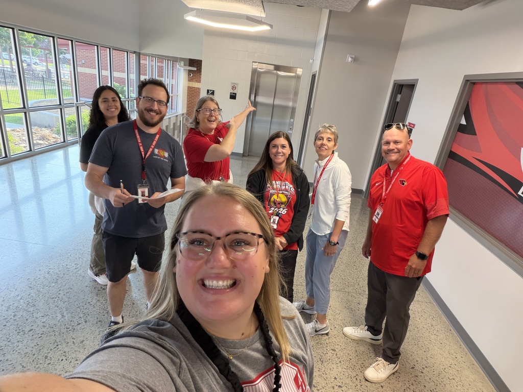 Group of seven people standing in a hallway with one person taking a selfie. Two large windows are behind them.