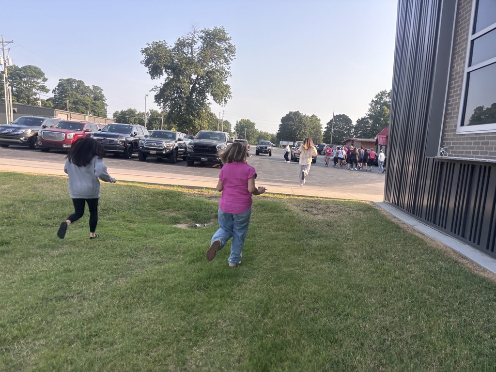 Two children run on a grassy field with parked cars and a building in the background.
