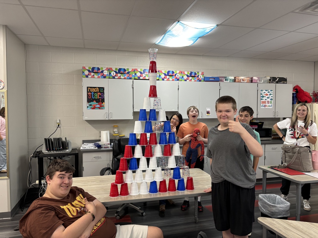 A group of students stands around a table with a pyramid of cups in a classroom.