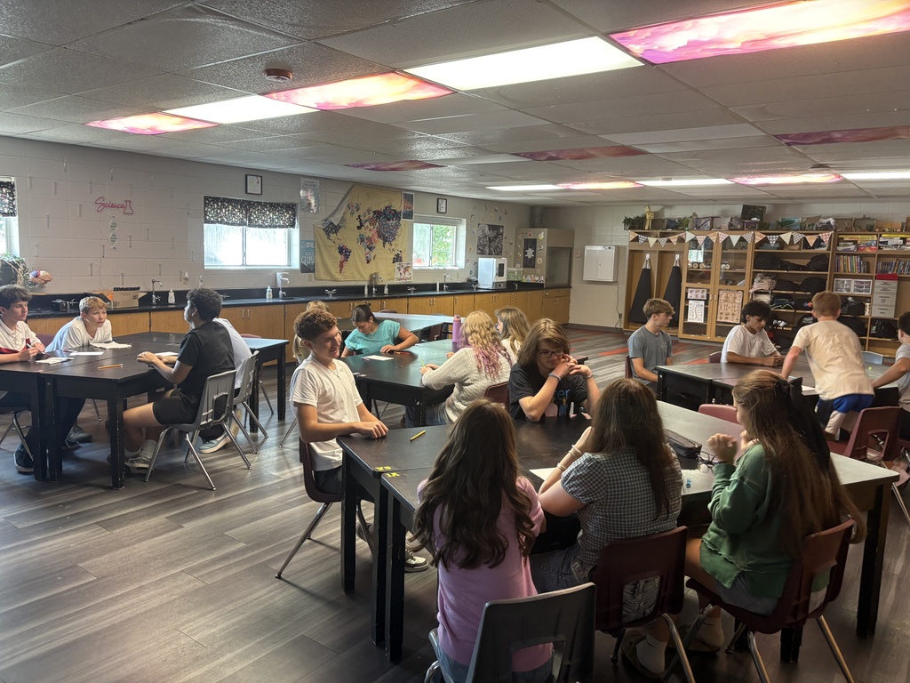 A classroom with wooden floors. Students in shirts sit at tables, facing each other, with a bookshelf in the background.