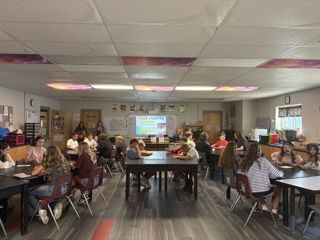 In a classroom, students sit at desks, some seated, some standing, listening to a teacher at the front.