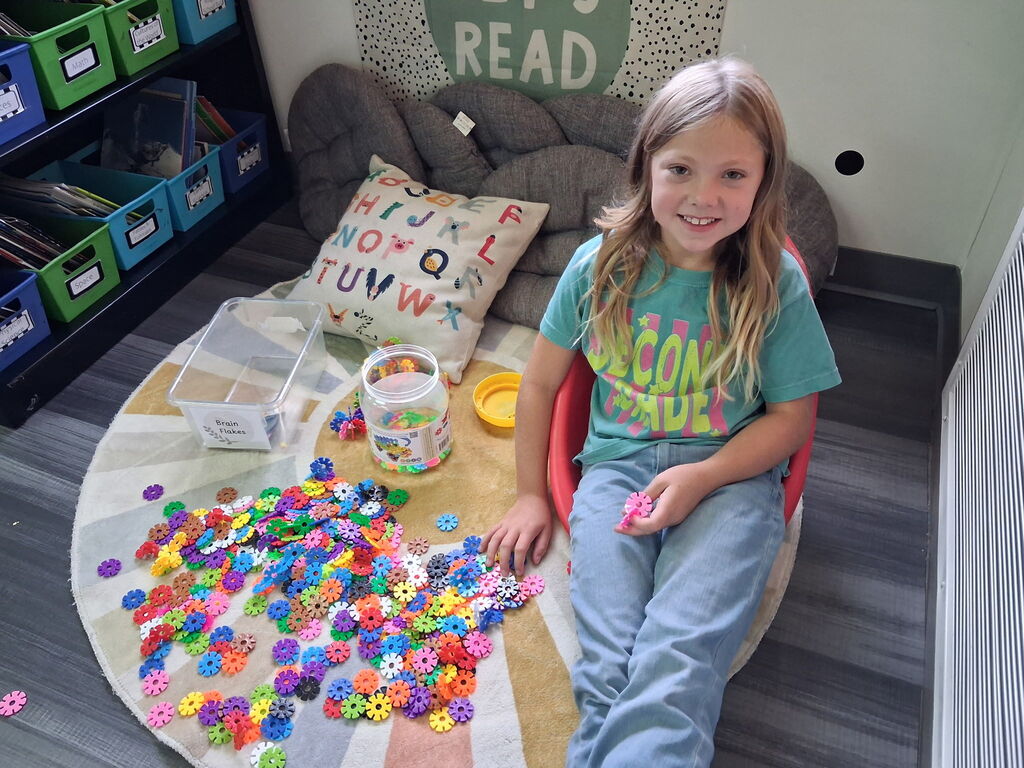 A child sits on a mat with a colorful array of buttons and beads. She has a book and a jar nearby.