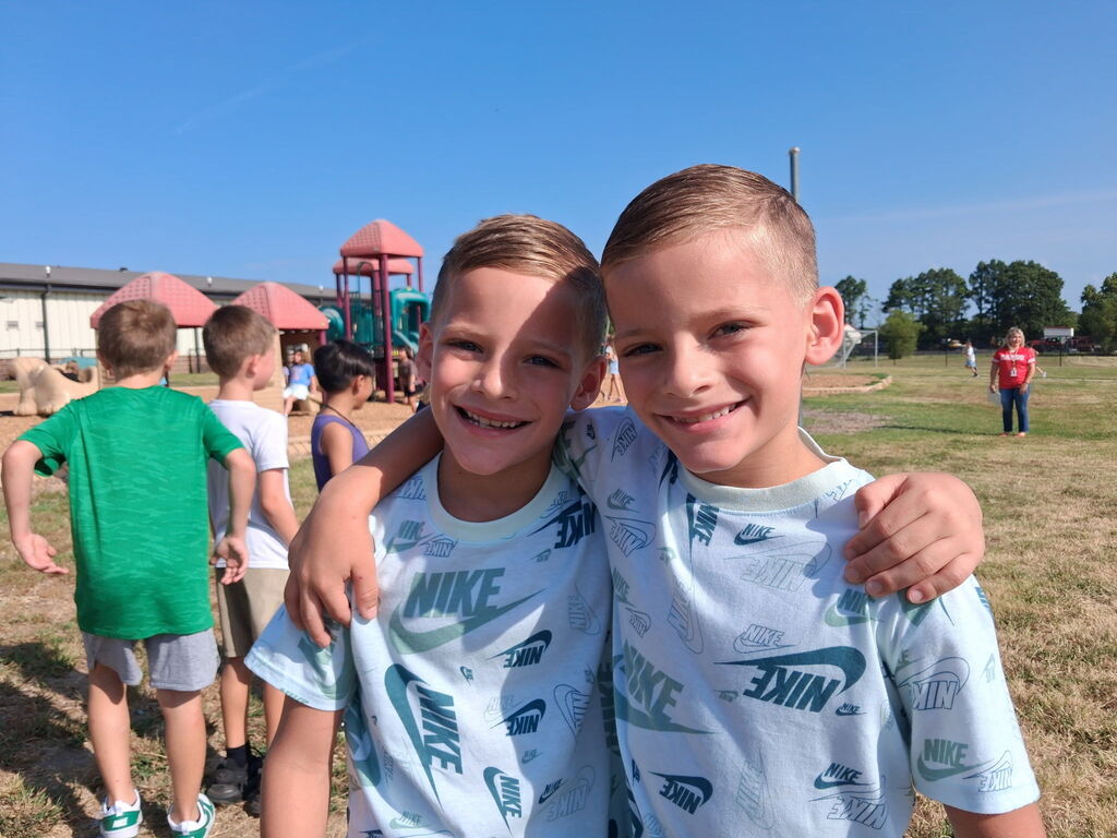 Two smiling boys stand together in a grassy field with playground equipment in the background.