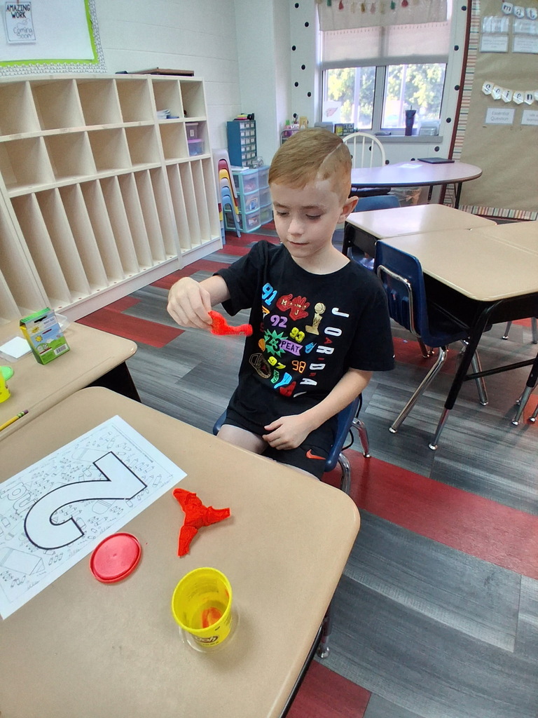 A child sits at a desk, holding a red object. A number 5 on paper and a yellow container are on the table.