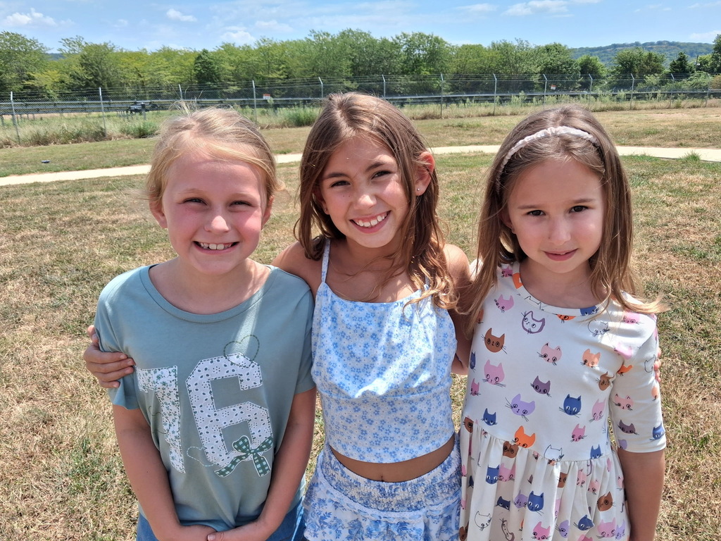 Three girls stand in a field. Two are smiling, one with arms around the others. Background has trees and grass.
