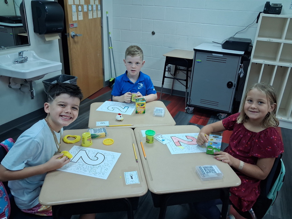 Three children seated at desks in a classroom, smiling. Each child has paper with numbers and art supplies.