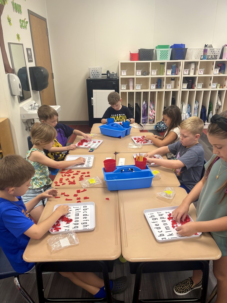 Kids seated at tables, some wearing masks, use red tiles on trays. A woman stands at the end of the table.