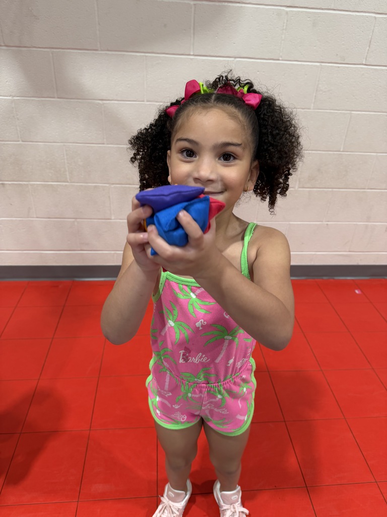 A young girl with curly hair holds a blue and purple ball in a gym setting.