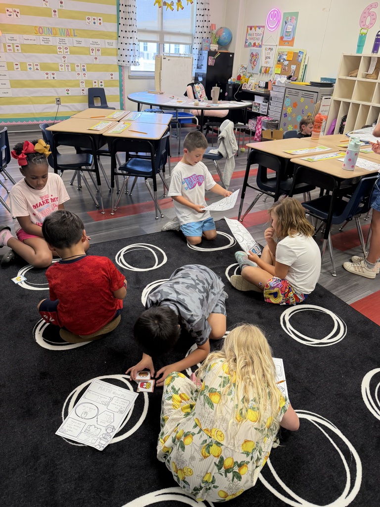 Children sit on a carpet with circles, some reading while others hold papers. Desks, chairs, and a globe are in the background.