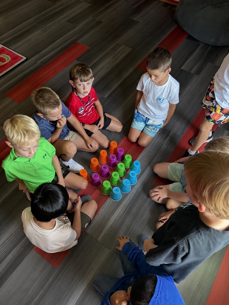 Nine children are sitting on the floor in a circle around a stack of colorful cups.