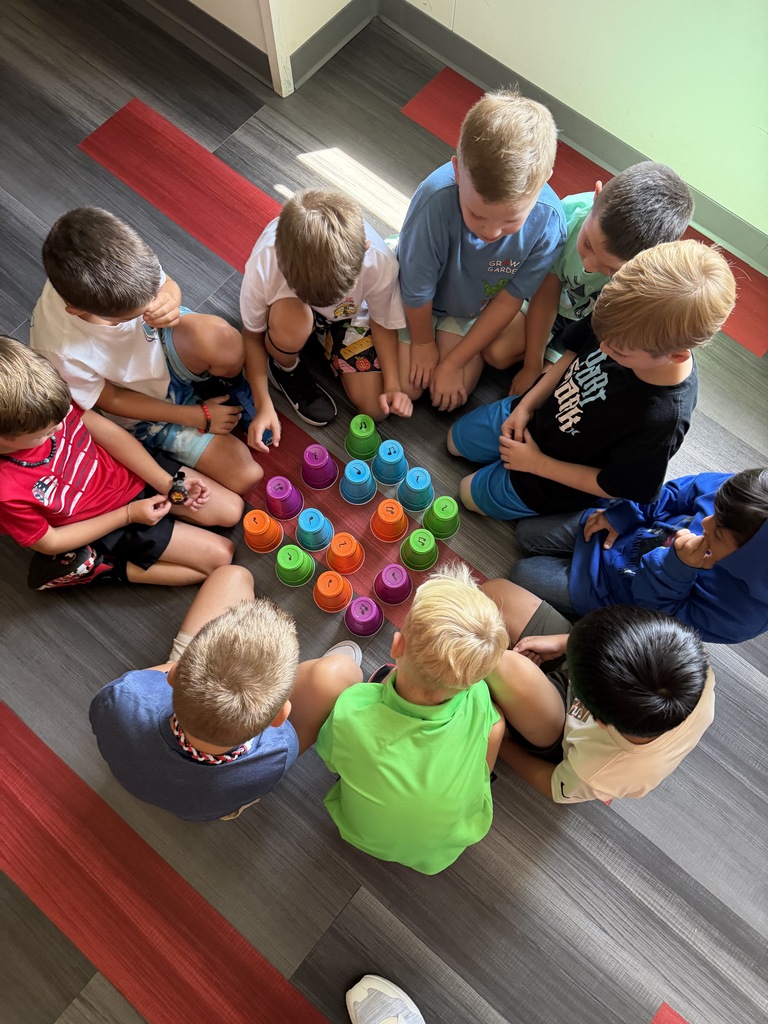 Several children are seated in a circle, playing with colorful cups on a wooden floor.