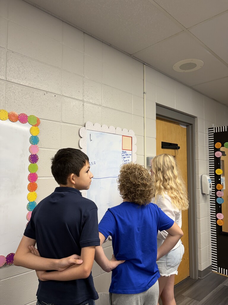 Three children stand near a wall, with two of them observing a bulletin board adorned with multicolored circles.