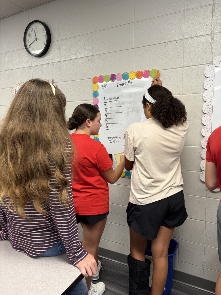 Four people stand near a bulletin board in a classroom. One person pins a paper.