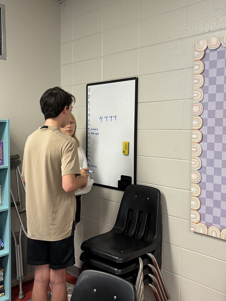 Two people standing by a whiteboard with numbers, one holding a pen, a white wall, and a black chair.