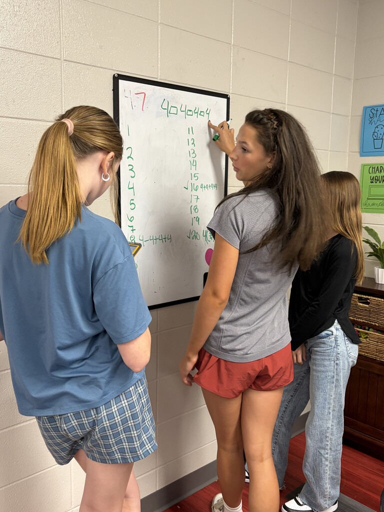 Three women work together at a wall. One writes on a whiteboard while the others watch.