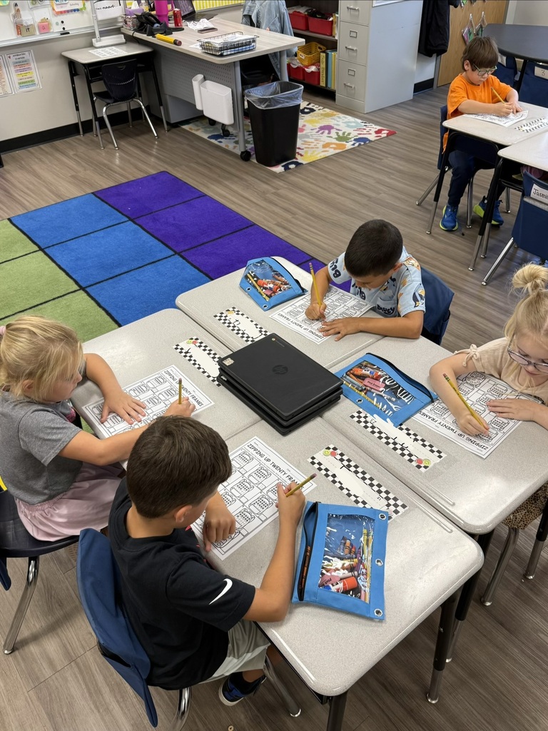 Children sitting at tables in a classroom. Some have binders, pencils, and worksheets in front of them.