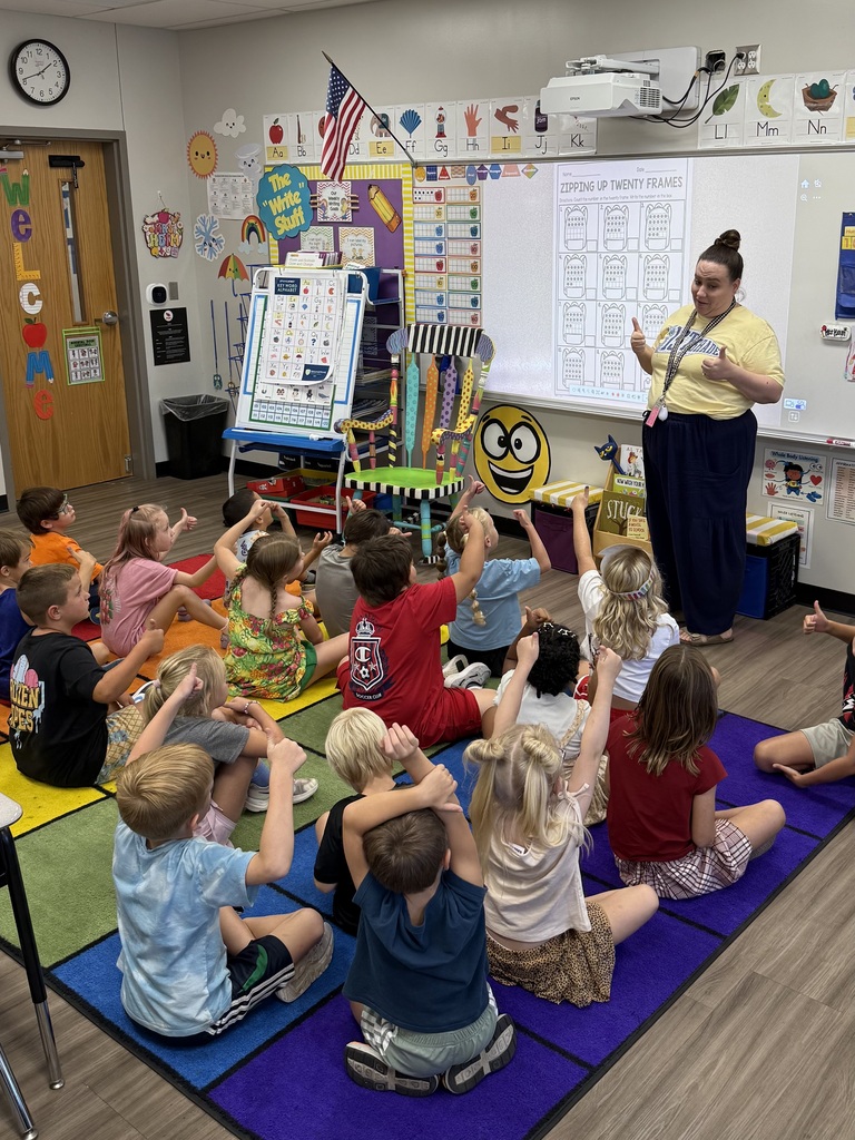 Teacher stands in front of class with projector screen. Children sit on rug and raise hands.