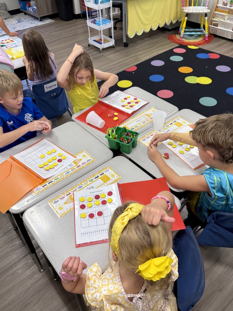 Four children seated at tables in a classroom with colorful floor mats, working on activities.