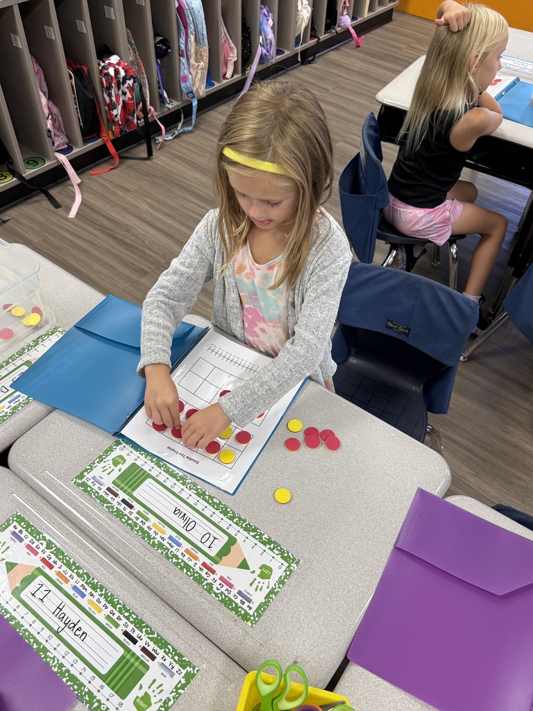 Child seated at a desk, using red and yellow tokens on a worksheet, with other students and school supplies around.