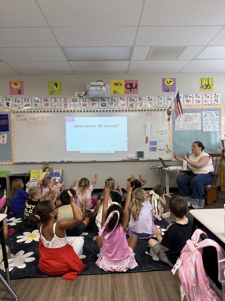 Children seated on floor, facing a teacher at a front table in a classroom. Whiteboard and projector screen visible.