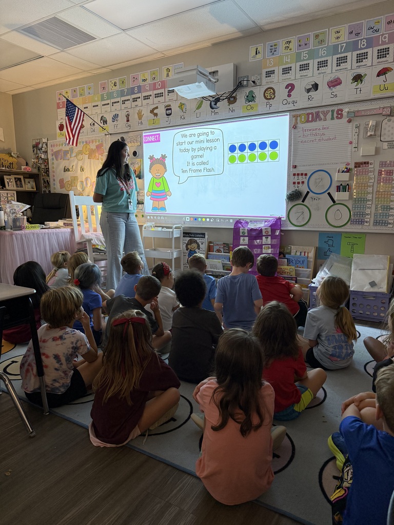 Woman teaching in front of screen, children sitting on floor in front of her, listening.