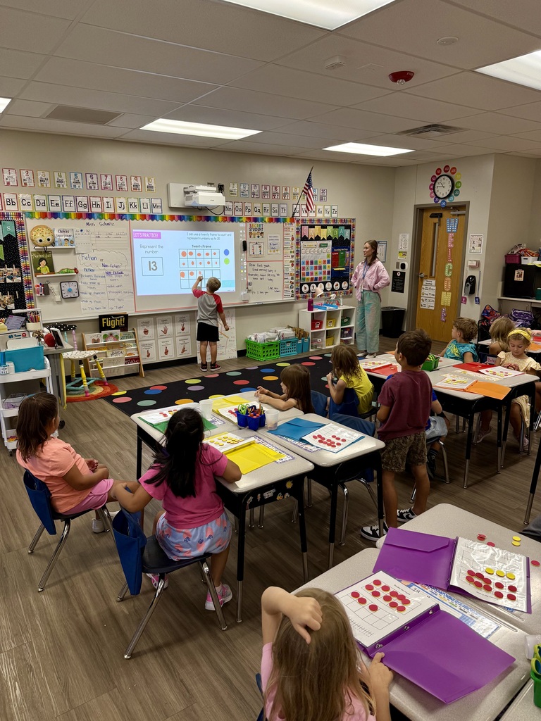 A classroom with a teacher at a whiteboard, children seated at desks, and a projector screen.