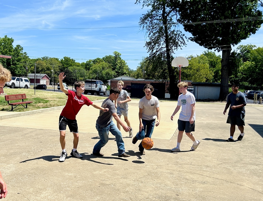 Several young people play basketball in a paved area with trees and a basketball hoop.