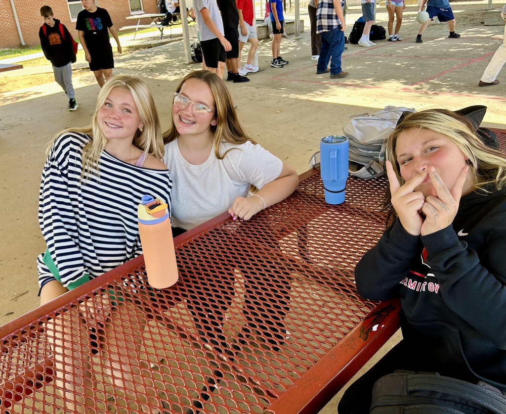 Three girls sit at a table with cups, posing for a photo, with a schoolyard in the background.