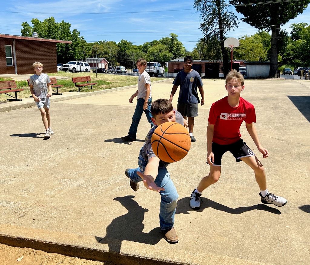 Five young men playing basketball in a paved area with two benches and trees in the background.