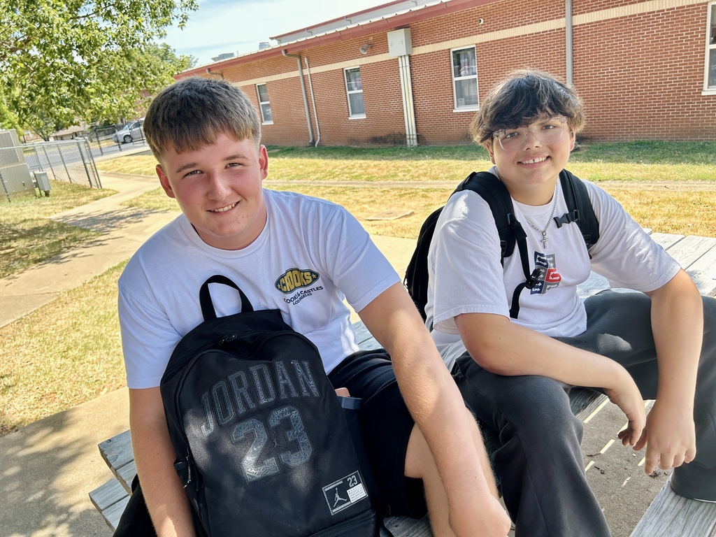 Two boys sit on a bench outside a brick building, wearing white t-shirts, glasses, and carrying backpacks.