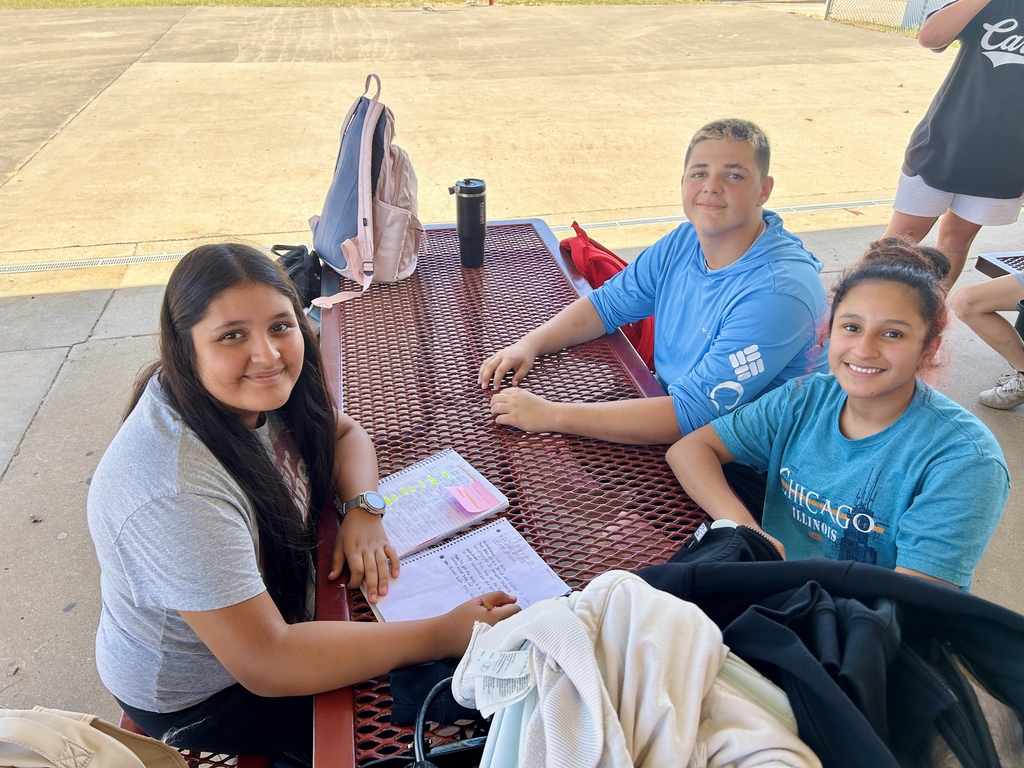 Three women sit at a table with papers. One wears a watch, another a backpack. Background shows an empty field.