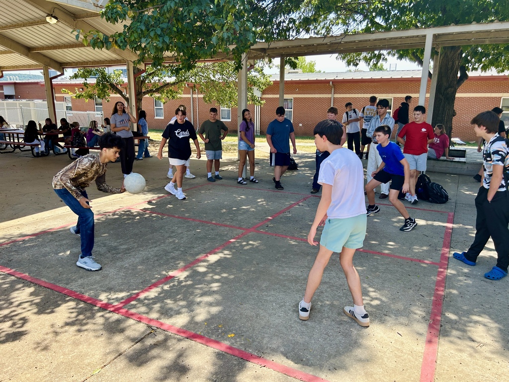 A group of children plays a ball game outside. The ground has red markings. Trees are in the background.