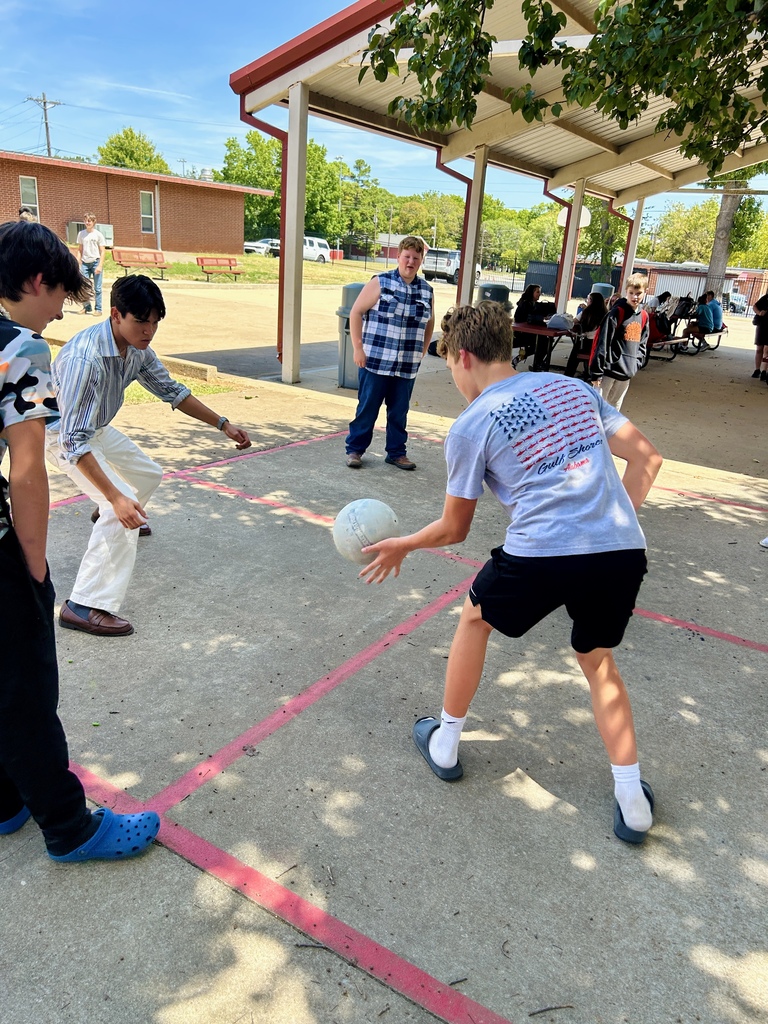 Four people play a game with a ball on a court with pink lines. A man watches in a plaza.